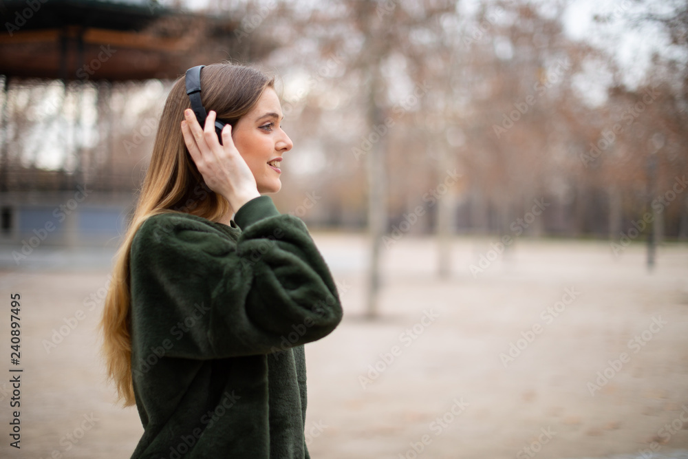 Side view of pretty young female in headphones smiling and listening to music while standing on blurred background of autumn park Side view of pretty young female in headphones smiling and listening to music while standing on blurred background of autumn park