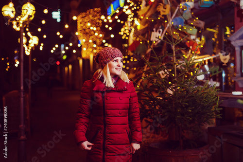 Outdoor photo of young beautiful happy smiling girl posing in street. Festive Christmas fair on background. Model wearing stylish winter coat, knitted beanie hat, scarf. © Angelov