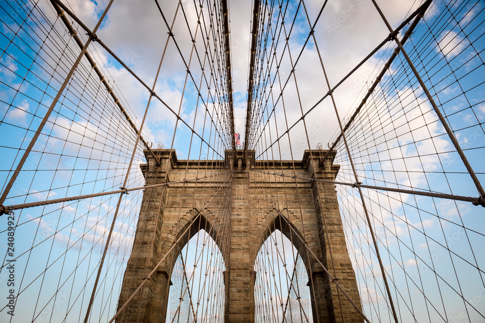 Fototapeta premium Classic view of the steel cable pattern and rough brick details of a stone tower of the Brooklyn Bridge under soft picturesque clouds