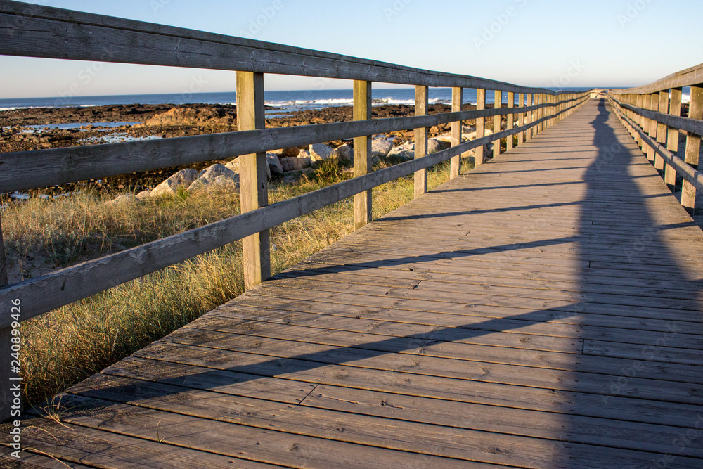 Obraz premium Wooden pier with grass on Atlantic ocean coast, Portugal. Wooden boardwalk to sea with rocks on shore. Empty walkway in dunes at dawn. Perspective background. Tropical travel concept. Peaceful place.