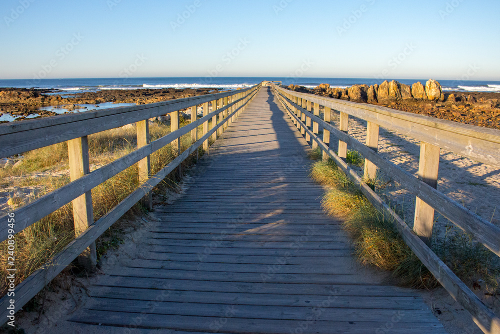Wooden pier with grass on Atlantic ocean coast, Portugal. Wooden boardwalk on sea with rocks on shore. Empty walkway in dunes at dawn. Perspective background. Tropical travel concept. Peaceful place.