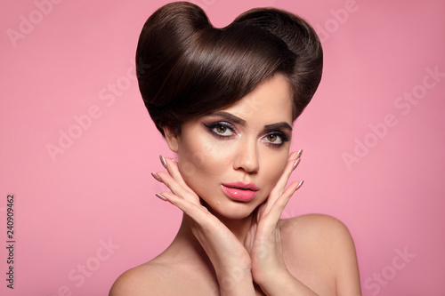 Glossy lipstick. Beauty portrait of High Fashion model with colorful bright makeup and pinup shiny hair style. Surprised brunette face holds cheeks by hand isolated on pink studio background.