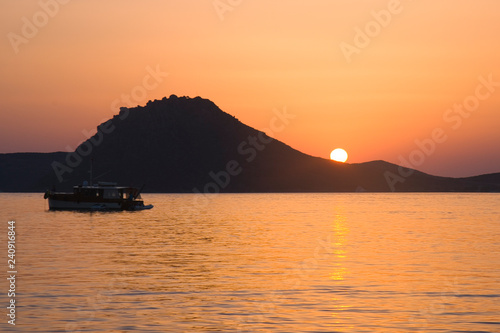 A sunset with sun falling below mountain on the horizon and calm waters in the foreground with boat sailing by.