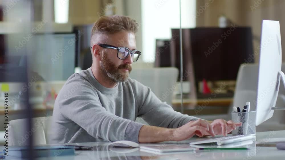 Medium shot of bearded young man in glasses sitting at office desk and typing on desktop computer keyboard Medium shot of bearded young man in glasses sitting at office desk and typing on desktop computer keyboard