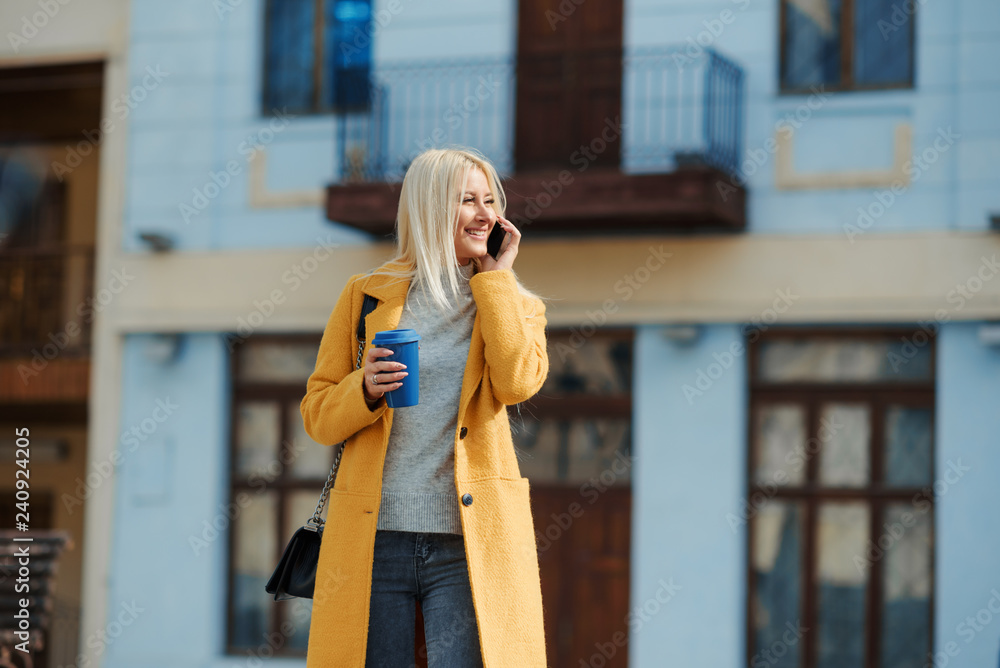 Fototapeta premium A young blond woman in a yellow bright coat is standing in the middle of the street and talking on the phone. Waiting for a meeting of friends. Holds a cup of coffee to go.