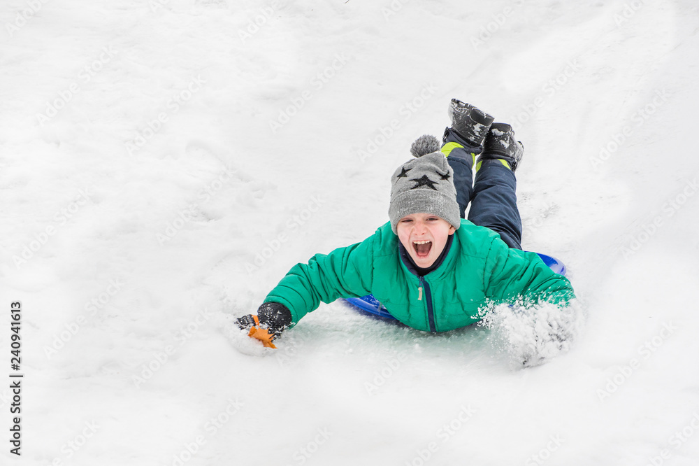 Laughing boy rides on his stomach from a hill on snow saucer. Seasonal concept. Winter day