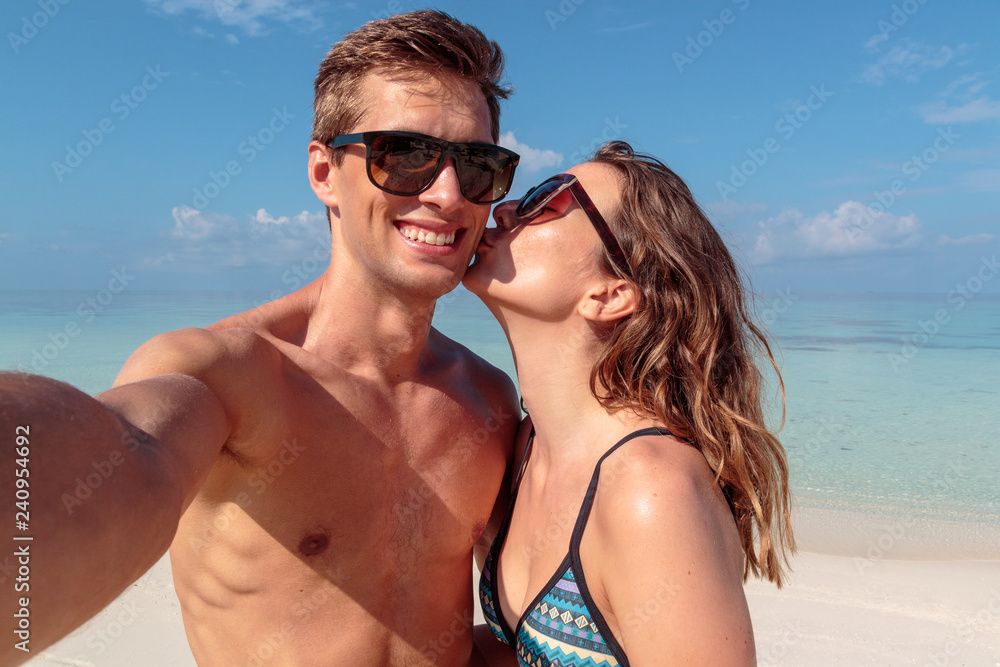 happy young couple taking a selfie, clear blue water as background. Girl kissing his boyfriend happy young couple taking a selfie, clear blue water as background. Girl kissing his boyfriend
