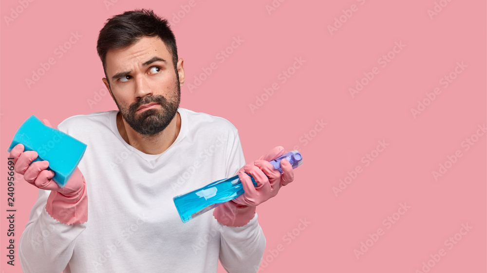 Serious hesitant man with thick bristle, looks confusingly, holds sponge and spray for cleaning glass surface, has thoughtful expression, stands over pink studio wall with blank space for your advert Serious hesitant man with thick bristle, looks confusingly, holds sponge and spray for cleaning glass surface, has thoughtful expression, stands over pink studio wall with blank space for your advert