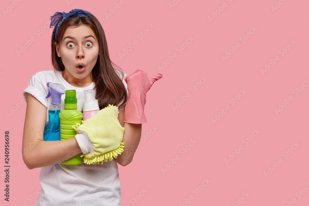 Scared emotive woman with surprised expression, wears headband, carries chemical detergents, indicates aside, shows dirty place for cleaning, keeps jaw dropped from fear. Housekeeping concept Scared emotive woman with surprised expression, wears headband, carries chemical detergents, indicates aside, shows dirty place for cleaning, keeps jaw dropped from fear. Housekeeping concept