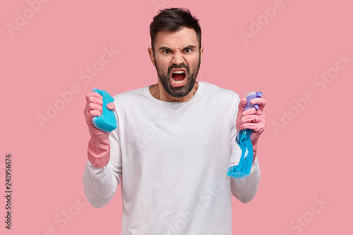 Studio shot of angry unshaven young man opens mouth from anger, holds spray and sponge, frowns face in discontent, expreses negative emotions, washes furniture, isolated over pink background