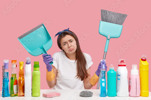 Dejected upset young housemaid frowns face in discontent, holds scoop and broom, does cleaning, wears headband and t shirt, isolated over pink background. Domestic routine and busyness concept