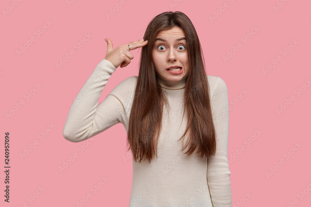 Photo of nervous puzzled Caucasain lady makes suicide gesture in temple, shoots in head, has displeased facial expression, wears casual white sweater, isolated over pink background. Body language Photo of nervous puzzled Caucasain lady makes suicide gesture in temple, shoots in head, has displeased facial expression, wears casual white sweater, isolated over pink background. Body language