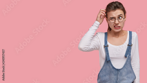 Photo of puzzled woman scratches head in bewilderment, thinks about finding right solution, wears trendy sarafan, round spectacles, models over pink background with free space for information photo