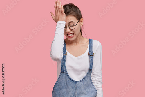 Photo of dissatisfied young woman regrets wrong doing, keeps hand on forehead, clenches teeth, dressed in fashionable outfit, isolated over pink studio wall. Lady forgets something important