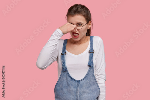 Obraz na plátně Unhappy dissatisfied woman frowns face, holds nose because of bad smell, clenches teeth from stinch, dressed in fashionable outfit, isolated over pink background
