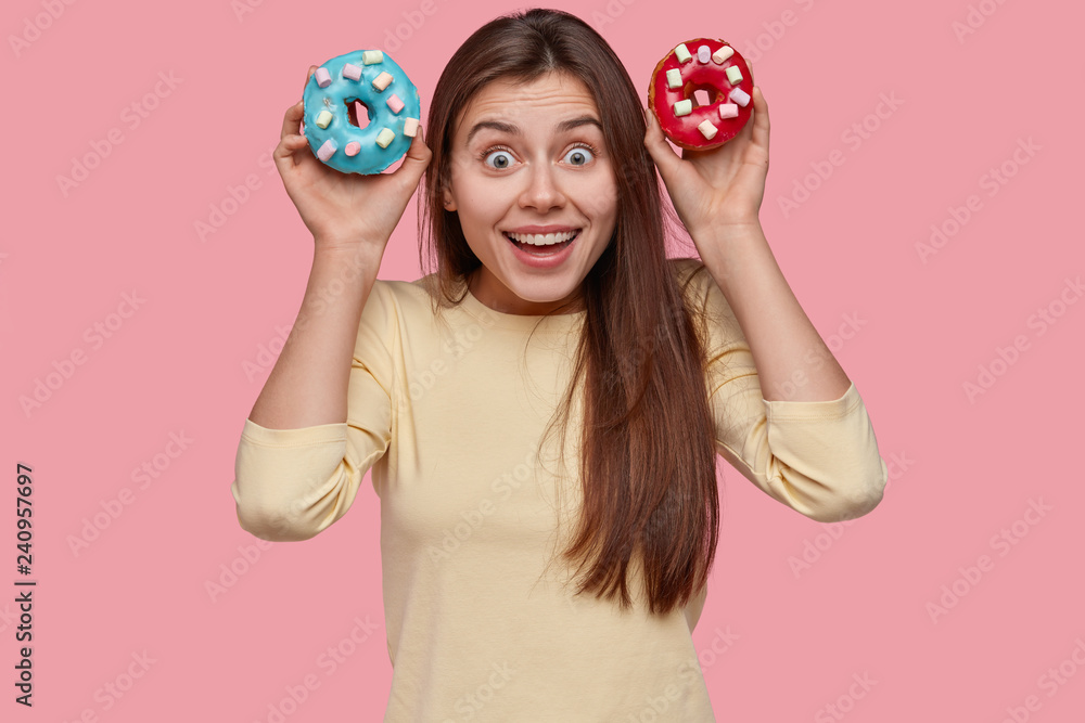 Funny brunette lady woman holds colourful doughnuts, looks happily directly at camera, dressed in casual clothes, models over pink wall. Junk food and nutrition concept. Positive woman with dessert Funny brunette lady woman holds colourful doughnuts, looks happily directly at camera, dressed in casual clothes, models over pink wall. Junk food and nutrition concept. Positive woman with dessert
