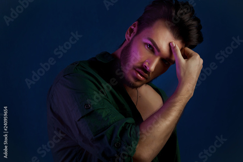 Fashion portrait of young man in green shirt and style haircut poses over dark blue wall with purple contrast color light