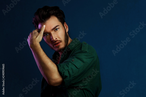 Fashion portrait of young man in green shirt and style haircut poses over dark blue wall with purple contrast color light