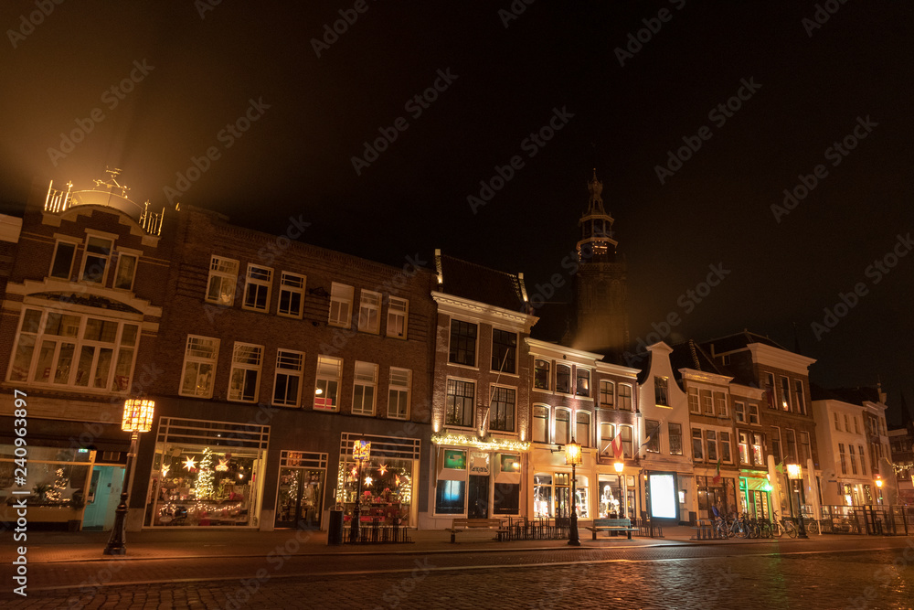Evening shot of a street with old houses with bell gables. In the background the tower of the Sint-Jans church with illuminated clock. Gouda, the Netherlands.