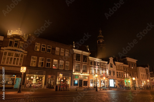 Evening shot of a street with old houses with bell gables. In the background the tower of the Sint-Jans church with illuminated clock. Gouda, the Netherlands.
