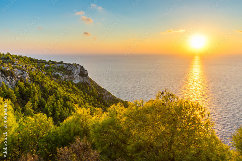Greece, Zakynthos, Orange sunset light over endless blue ocean behind green rough cliffs