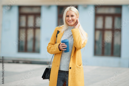 Coffee to go. Beautiful young blond woman in bright yellow coat holding coffee cup and smiling while walking along the street 