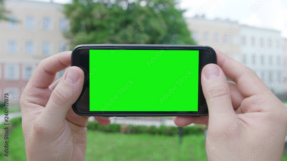 Lublin, Poland - July 2018: Two hands holding a phone with a green screen in a horizontal position in a public park. Close-up shot. Soft focus