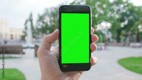 Lublin, Poland - July 2018: A hand holding a phone with a green screen in a public park. Close-up shot. Soft focus