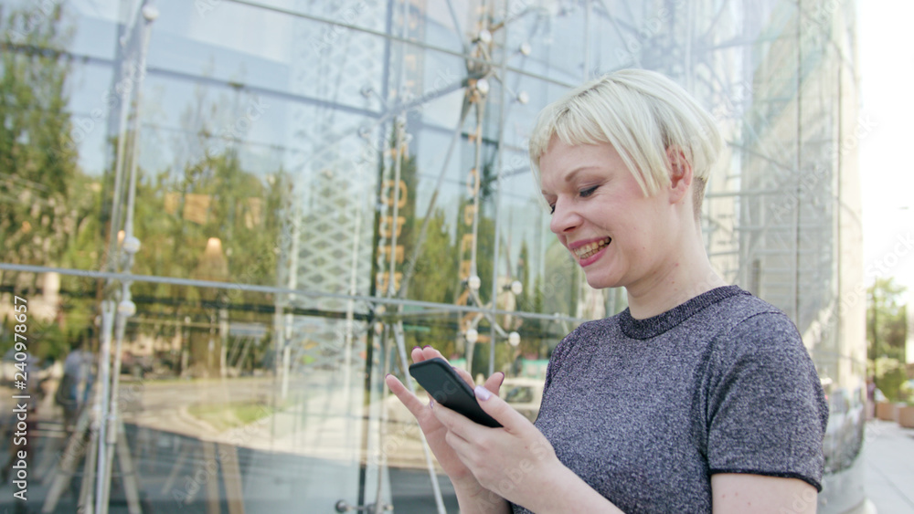 An attractive young blonde lady using a phone in town. Medium dolly shot. Soft focus