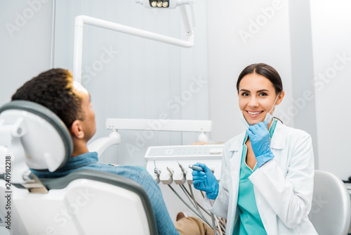 cheerful dentist holding drill and smiling near african american patient