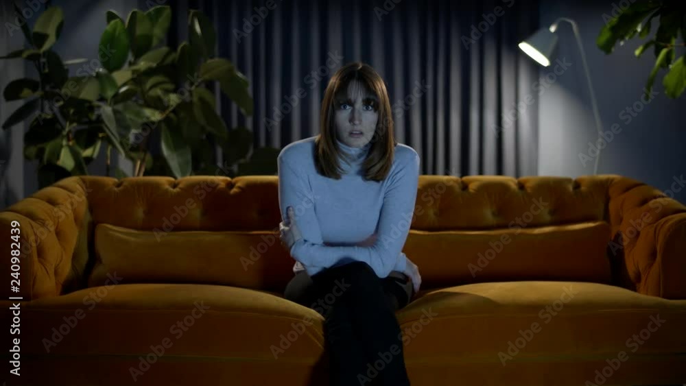 Young woman sitting on a cozy sofa at home at night watching tv reacts to something on screen with a shocked and surprised expression on her face. Young woman sitting on a cozy sofa at home at night watching tv reacts to something on screen with a shocked and surprised expression on her face.