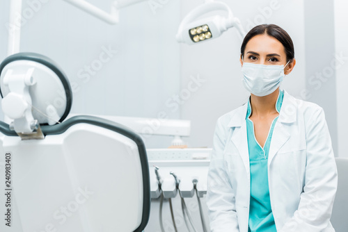 female dentist in white coat and mask near dental chair
