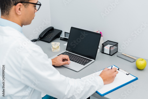 selective focus of male dentist making notes and using laptop with blank screen