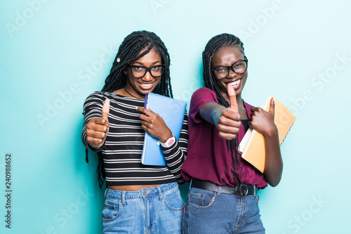 Portrait of two african young school teenage girls holding copybooks and showing thumbs up isolated over blue background
