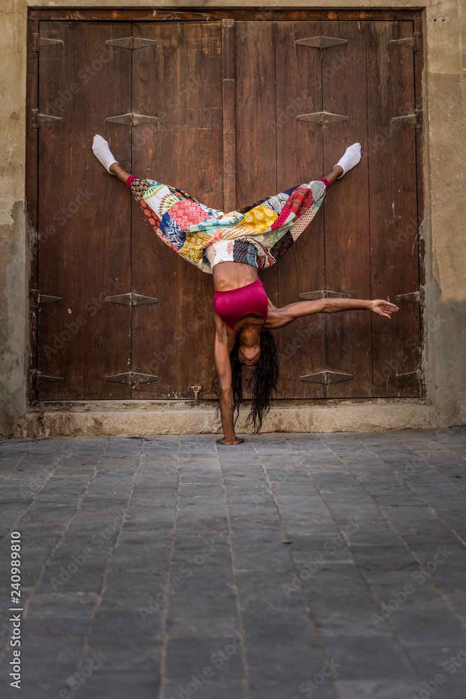 Beautiful tall athletic African American Woman does a one handed handstand  pose wearing a bright pink sports bra and a multicolored pant against an  old wooden door Stock Photo