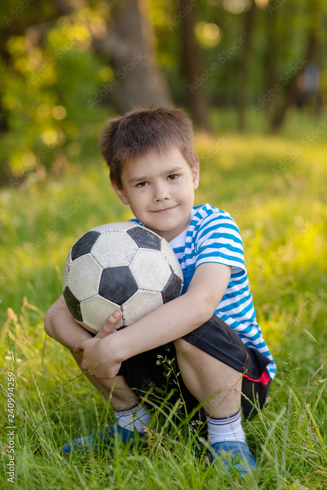 I love football. Joyful dark-haired boy smiling and sitting with his soccer ball