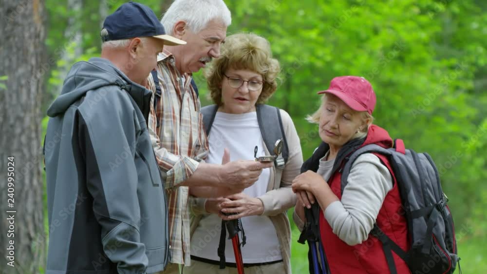 Tilt up shot of senior people with backpacks and trekking poles standing in forest and navigating with compass during hike Tilt up shot of senior people with backpacks and trekking poles standing in forest and navigating with compass during hike