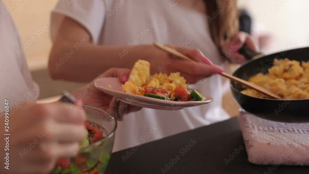 Two girls impose an omelet and vegetable salad of tomatoes and cucumbers in a plate, close-up. Two young girls making breakfast at home in the kitchen. Two girls impose an omelet and vegetable salad of tomatoes and cucumbers in a plate, close-up. Two young girls making breakfast at home in the kitchen.