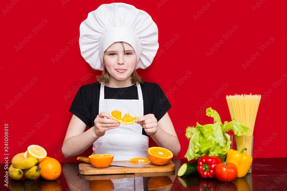 Happy little girl in chef uniform cuts fruit in kitchen. Kid chef. Cooking Concept Happy little girl in chef uniform cuts fruit in kitchen. Kid chef. Cooking Concept