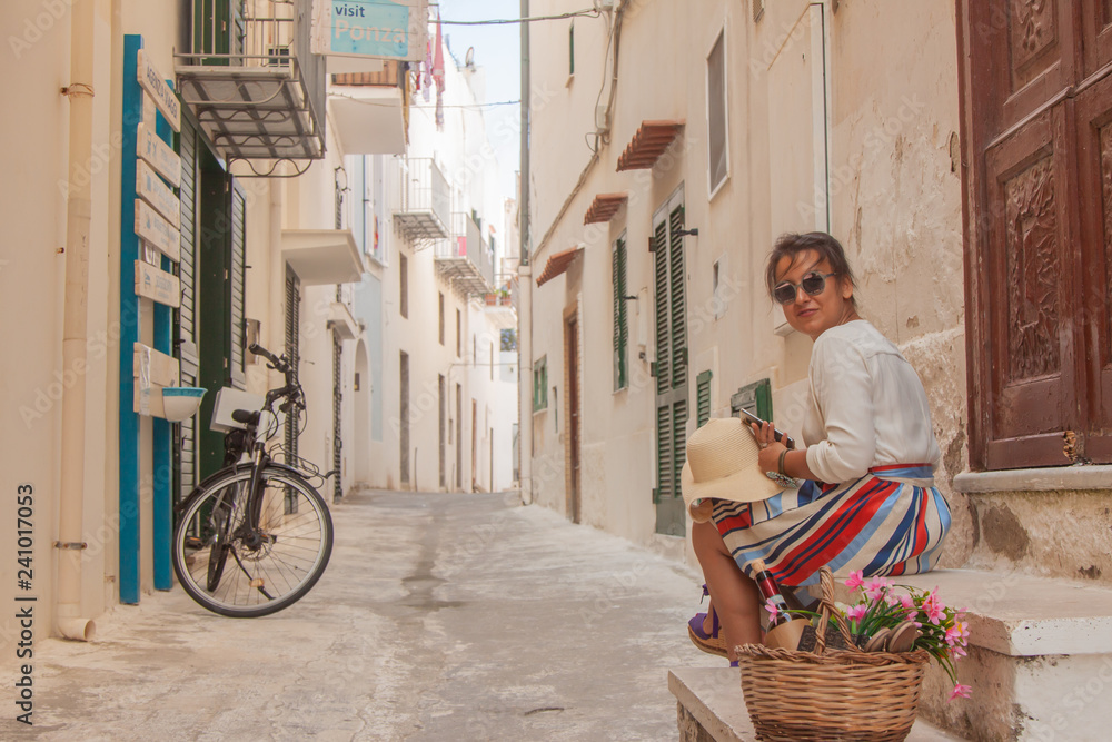 Young woman sitting on door steps in a typical alley on Ponza island town, Italy. Fashion dress and sunglasses, white building.