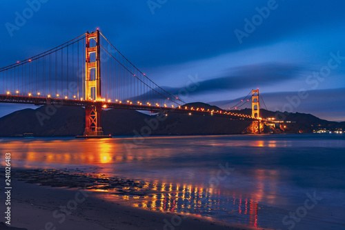 Fototapeta Naklejka Na Ścianę i Meble -  Nighttime view of Golden Gate Bridge reflected in the blurred water surface of San Francisco bay, dark blue sky background; California