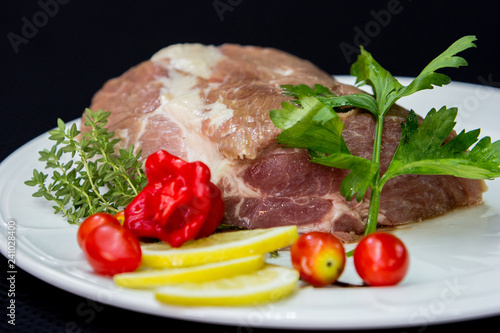 Raw uncooked buffalo meat prepared to cook in a white plate with tomatoes, lemon and herbs against a black background. (ID: 241028400)