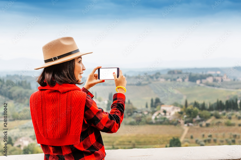 Young woman traveler with hat taking selfie with smartphone near famous Val Di Orcia valley, Tuscany, Italy