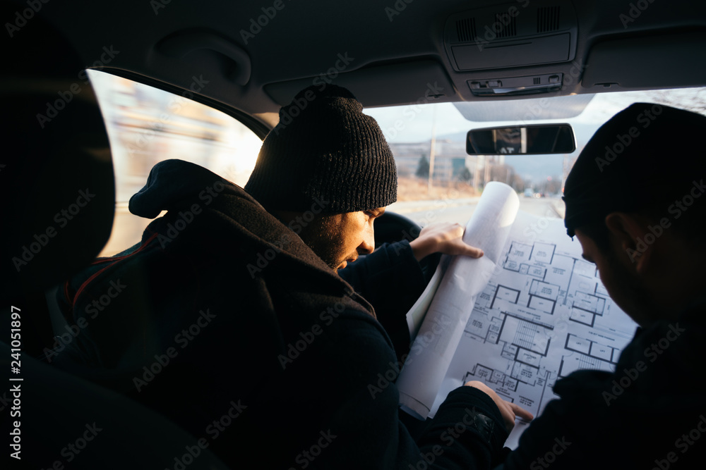 Two violent robbers sitting in a car looking at a blueprint of the building they want to rob while proudly showing off their guns.