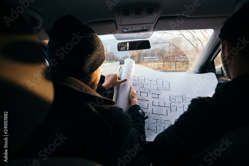 Two violent robbers sitting in a car looking at a blueprint of the building they want to rob while proudly showing off their guns.