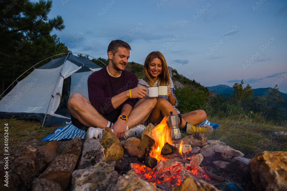 Young couple drinking coffee,  and warming up on the bonfire in the forest hill in the dusk