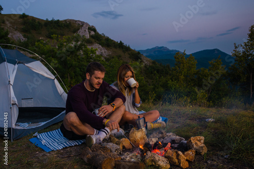 Young couple relaxing by the campfire and drinking coffee in the forest hill in the dusk