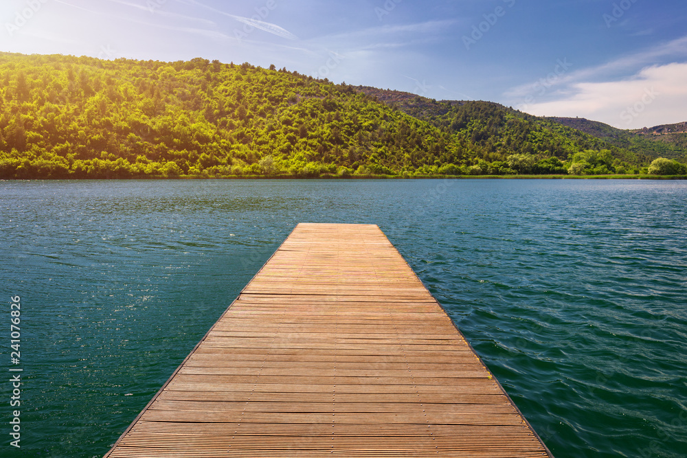 Idyllic view of the wooden pier in the lake with mountain scenery background. Wooden bridge on the lake. Wooden pier jutting out into the lake. Long pier on lake and blue sky in summer.