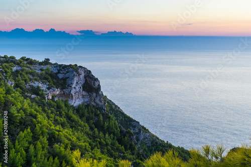 Greece, Zakynthos, Endless silent blue ocean behind green tree covered chalk cliff in evening twilight