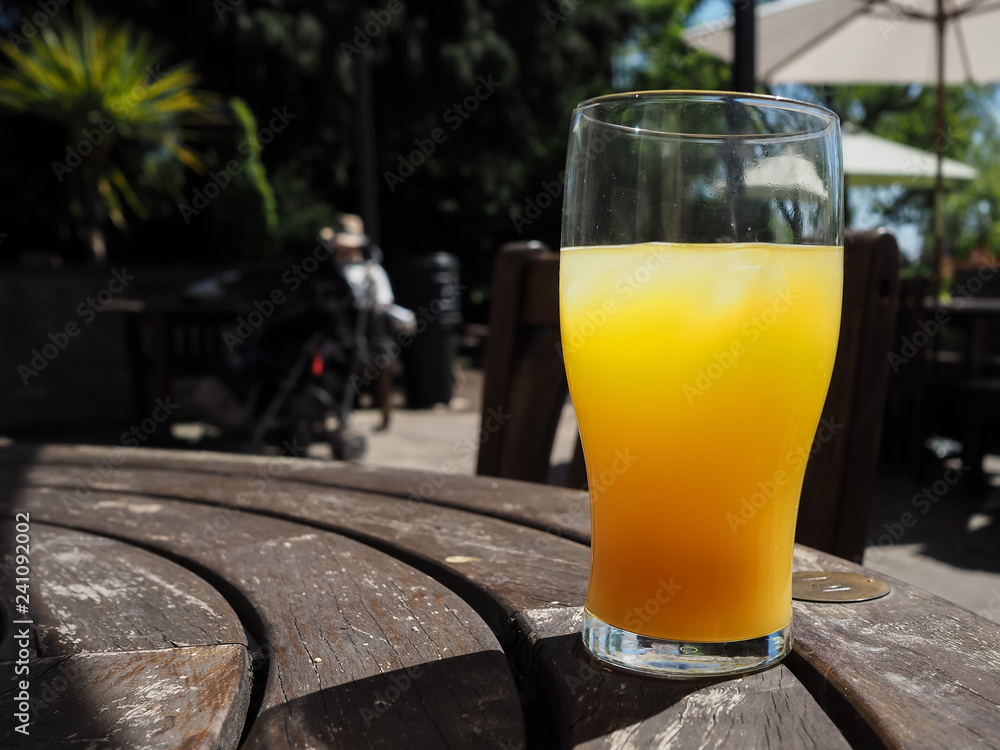 Pint glass full of orange juice on a hot day, on a wooden table with people blurred in the background